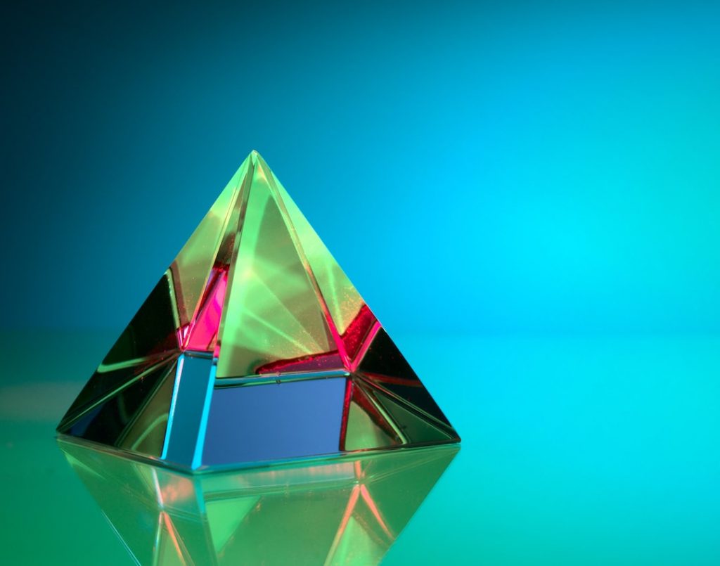 A glass prism shaped like a pyramid reflects and refracts colourful light, creating green, red, and yellow hues, set against a vibrant blue teal background, with reflections visible on the smooth surface below.
