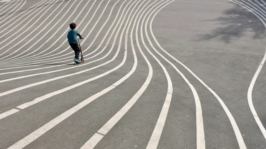 A child rides a scooter along a paved surface with curving white lines, creating a visually dynamic pattern. The lines curve and intersect, adding a sense of motion and depth to the scene.