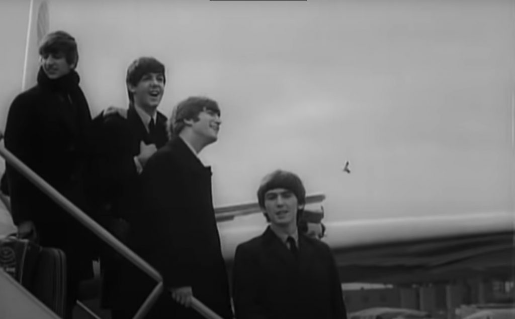Four young men in dark suits and coats stand smiling on an aeroplane staircase, one holding a suitcase, as they arrive for the Beatles’ United States tour under an overcast sky. Part of the aeroplane is visible behind them in New York.