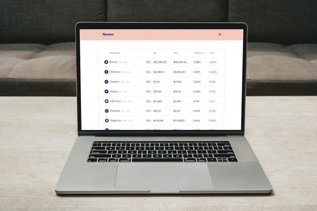 A laptop on a table displays a cryptocurrency trading platform, showing various coins like Bitcoin, Ethereum, and Cardano, along with their buy and sell prices, percentage changes, and market data.