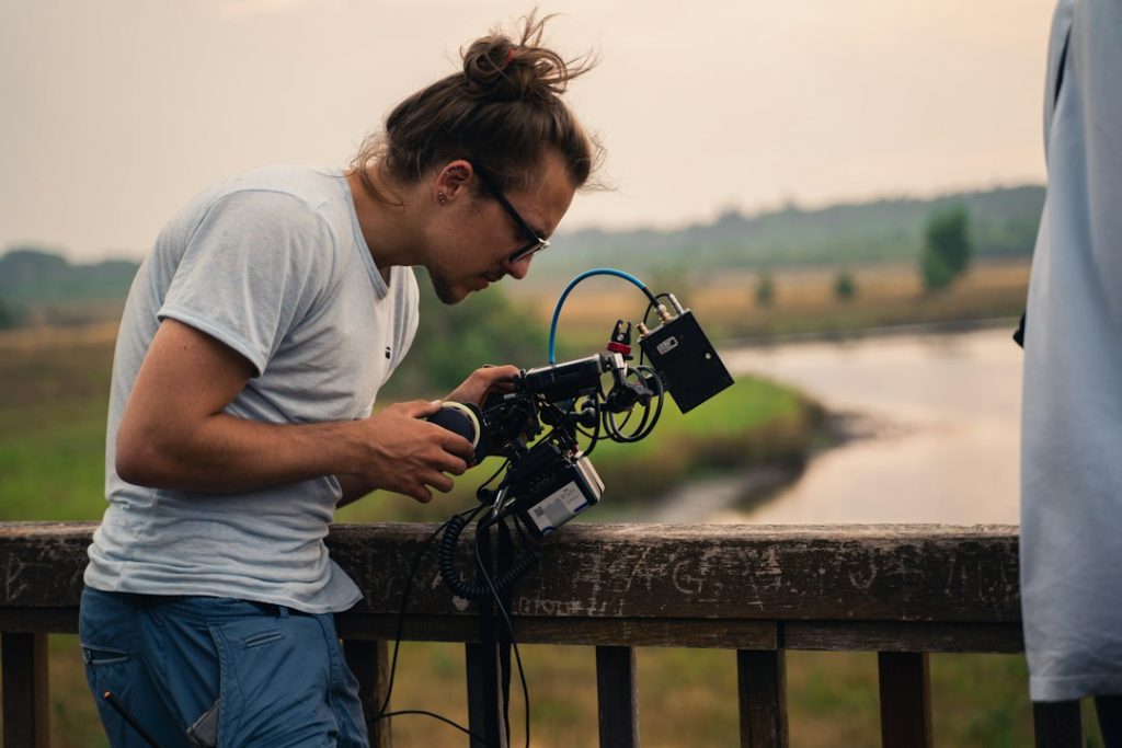A person with a bun and glasses leans over a wooden railing, intently adjusting a professional video camera, with a river and grassy landscape blurred in the background.