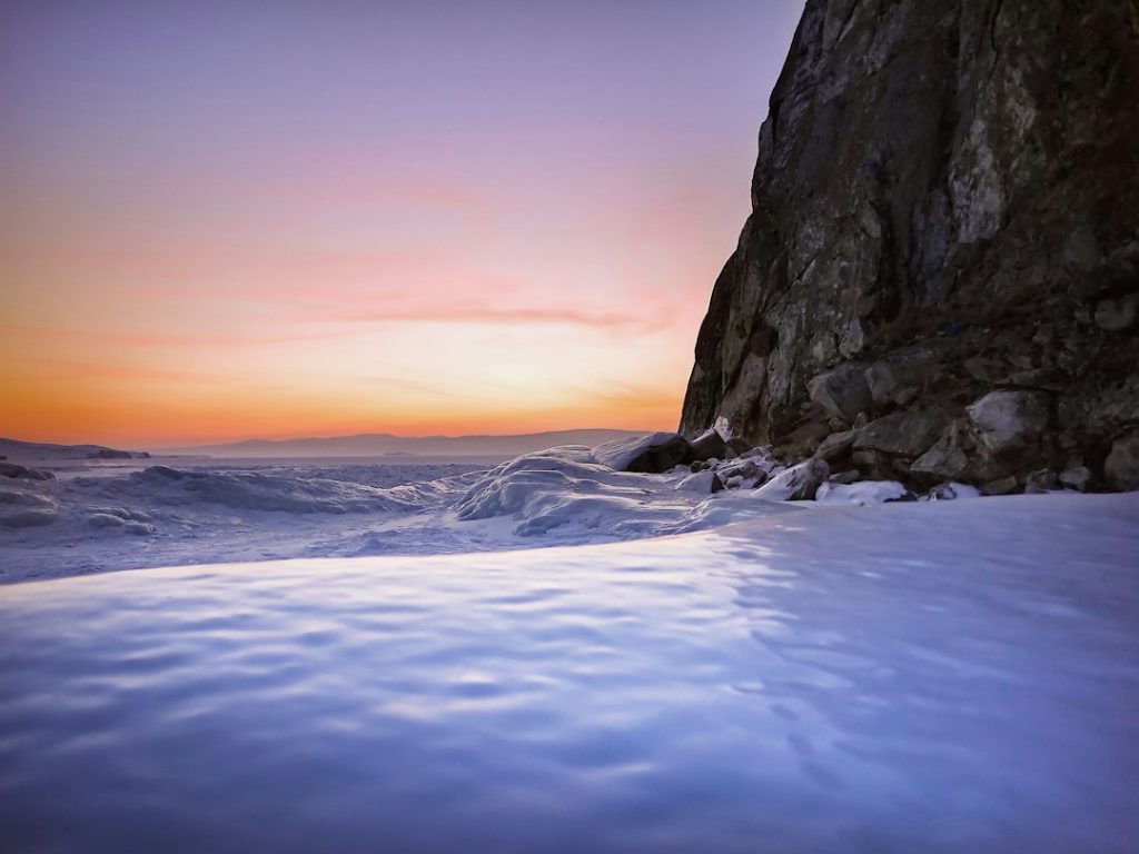 A serene winter landscape at sunset, featuring a snow-covered frozen lake, a rocky cliff on the right, and a colourful sky with soft hues of orange, pink, and purple in the background.