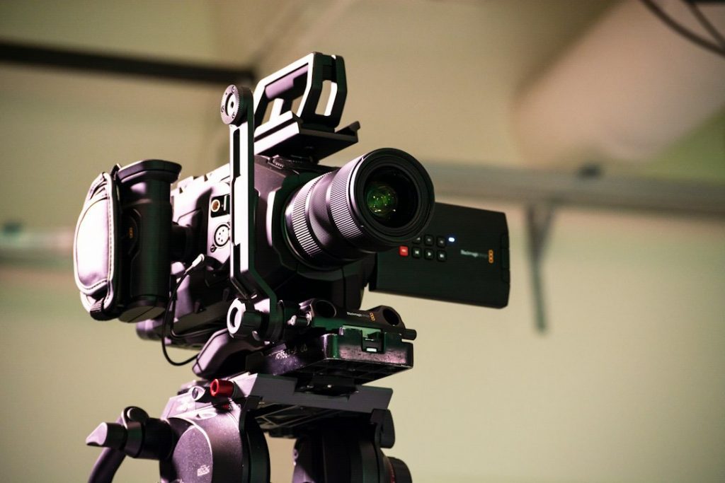 A professional digital video camera with a large lens is mounted on a tripod in an indoor setting, ready for filming.