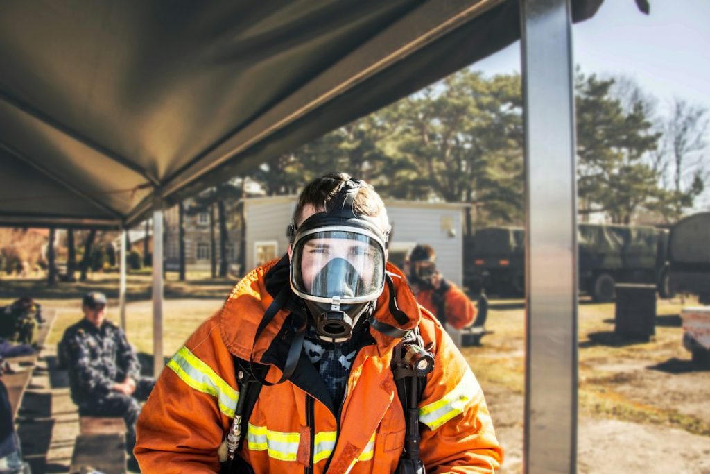 A firefighter wearing an orange suit and a full-face respirator mask stands under a canopy outdoors, with other people and emergency vehicles in the background on a sunny day.