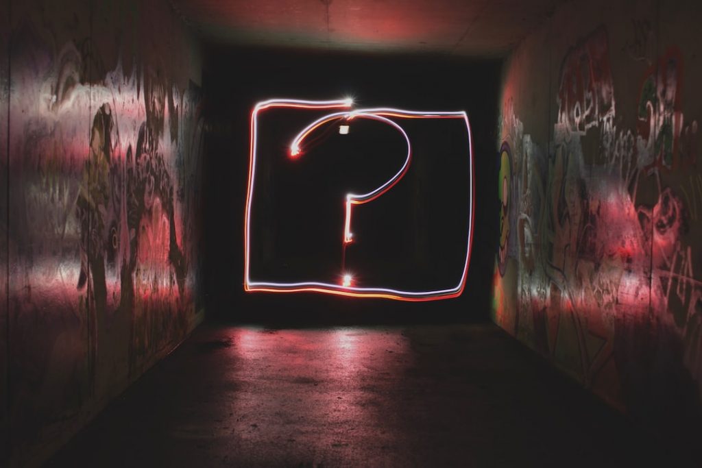 A glowing question mark drawn with light stands in the centre of a dark, graffiti-covered tunnel, creating a mysterious and dramatic atmosphere.