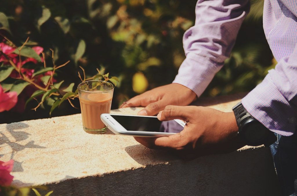 A person in a purple shirt is holding a mobile phone while standing outdoors next to a glass of tea placed on a ledge, with greenery and pink flowers in the background.