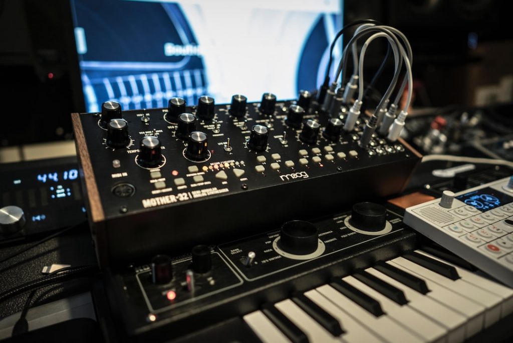 A close-up of electronic music equipment, including a Moog Mother-32 synthesiser with patch cables connected, resting on a small keyboard and surrounded by other audio devices in a studio setting.