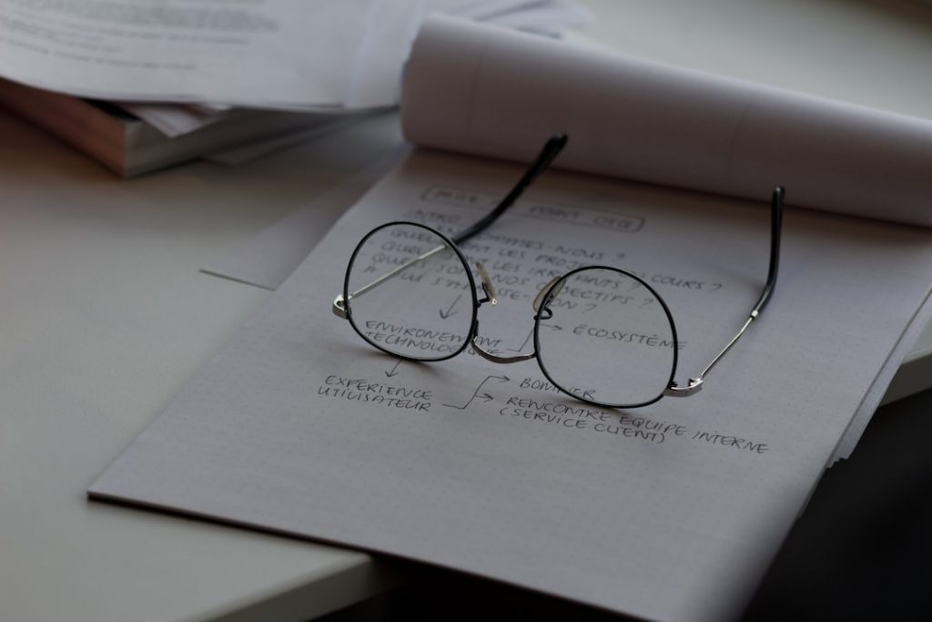 A pair of spectacles rests on a notepad with handwritten notes and a flowchart diagram. Papers are stacked in the background on a desk, lit by natural light.