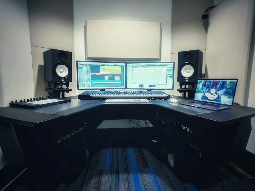 A modern music production studio setup with two monitors displaying digital audio workstations, speakers on each side, a keyboard controller, and a laptop showing an anime character, all on a black desk with equipment underneath.