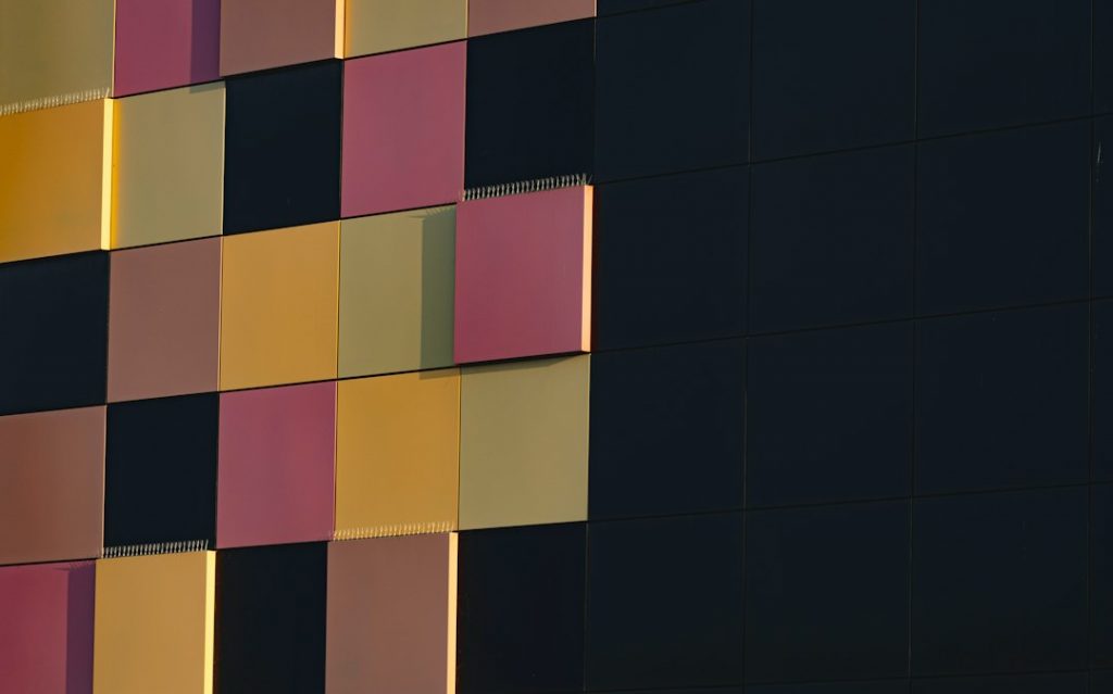 A building façade with a geometric pattern of square and rectangular panels in black, yellow, and pink hues, arranged in a grid with some panels projecting slightly.