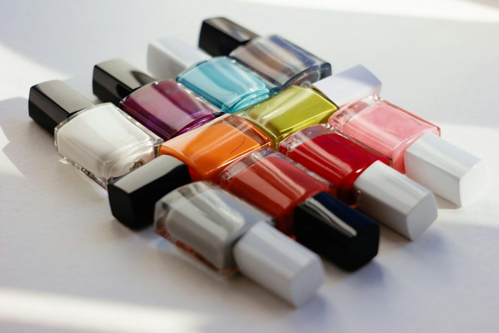 Ten bottles of nail varnish in various colours, including white, pink, orange, red, blue, yellow, grey, and purple, are arranged in a staggered pattern on a white surface with natural light.