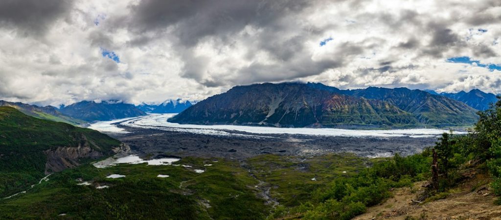 A panoramic view of a glacier winding through a lush green valley, surrounded by rugged mountains under a dramatic, cloudy sky.