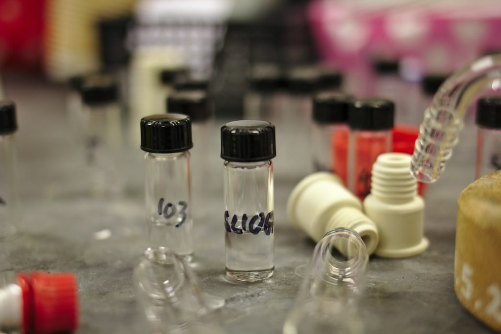 Several small glass vials with black caps and handwritten labels stand on a laboratory table, surrounded by other lab equipment and blurred background objects.