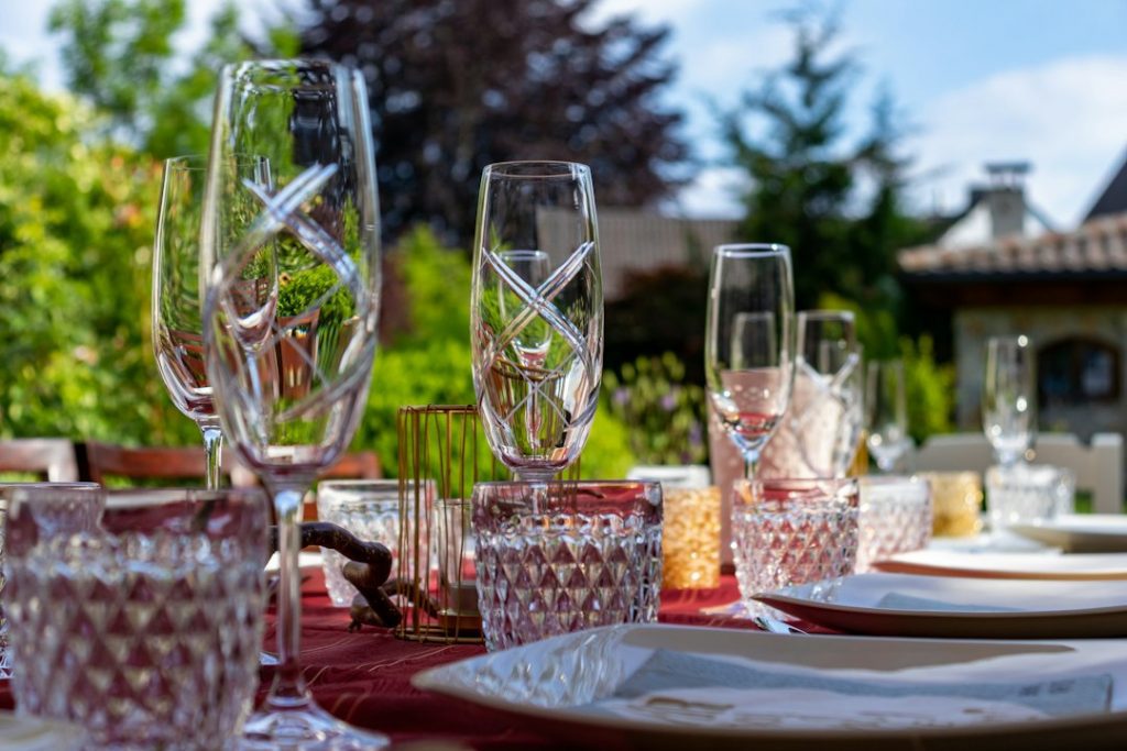 A close-up of an outdoor dining table set with crystal glasses, plates, and serviettes on a red tablecloth, with a garden and house blurred in the background on a sunny day.