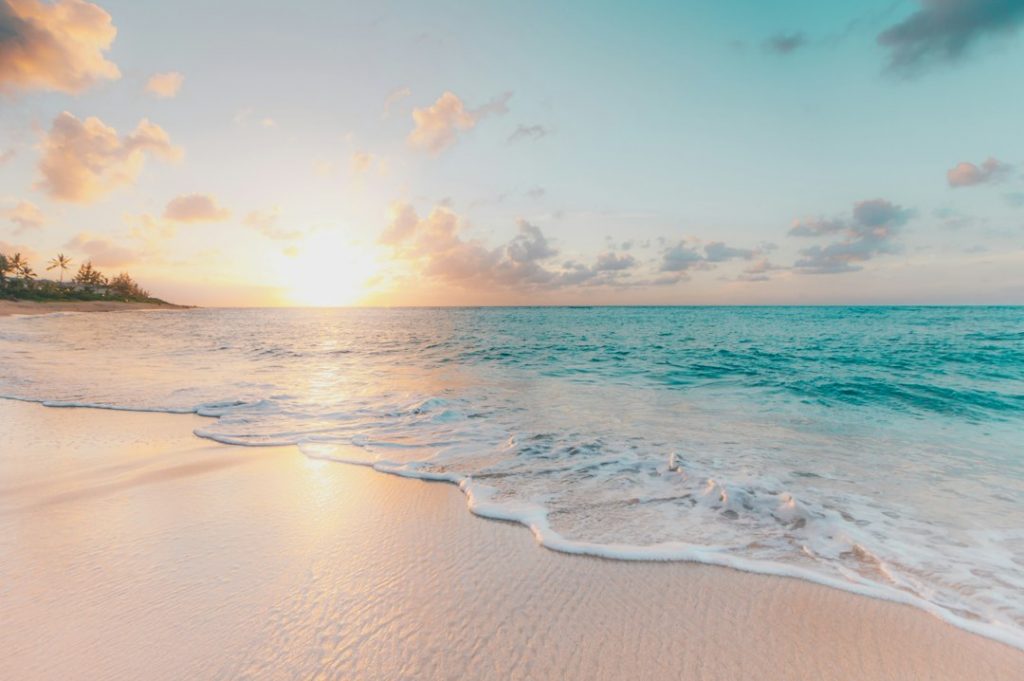 A serene beach scene at sunrise or sunset, with gentle waves washing onto soft sand and the sun glowing near the horizon. Light clouds are scattered in a pastel-coloured sky. Palm trees are visible in the distance.