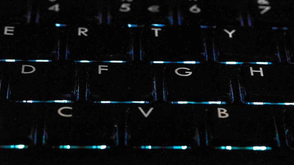 Close-up of a computer keyboard in dim lighting, showing the letters E, R, T, D, F, G, C, V and B, with blue light shining between the keys.