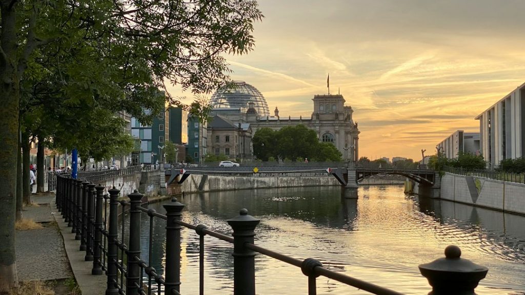 A peaceful canal in Berlin at sunset, with the glass dome of the Reichstag building in the background, trees lining the walkway, and a small bridge crossing the water.