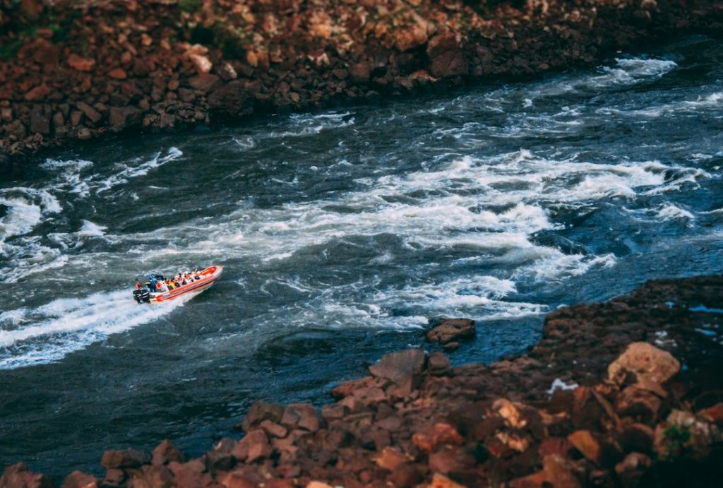 A small inflatable dinghy with several people navigates through fast-moving white-water rapids, surrounded by rocky riverbanks.
