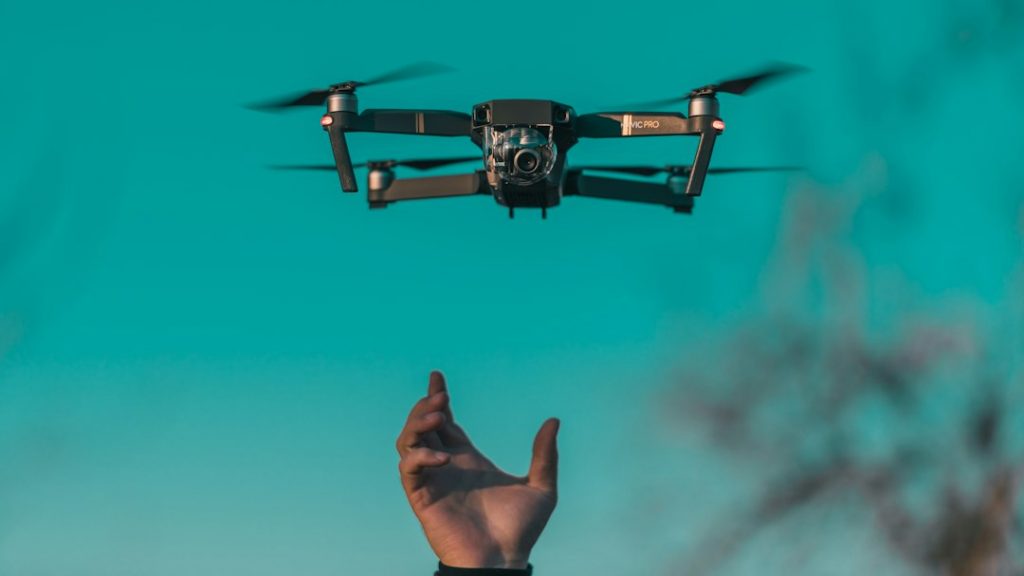 A person’s hand reaches up towards a flying drone hovering in the air against a clear teal sky. The drone’s camera faces forward, and its propellers are spinning.