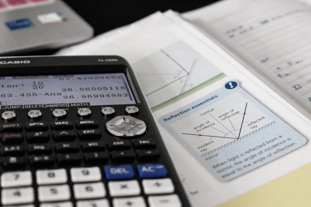 A scientific calculator rests on open maths and science textbooks, showing diagrams and calculations about light reflection. Handwritten notes and a laptop are partially visible in the background.