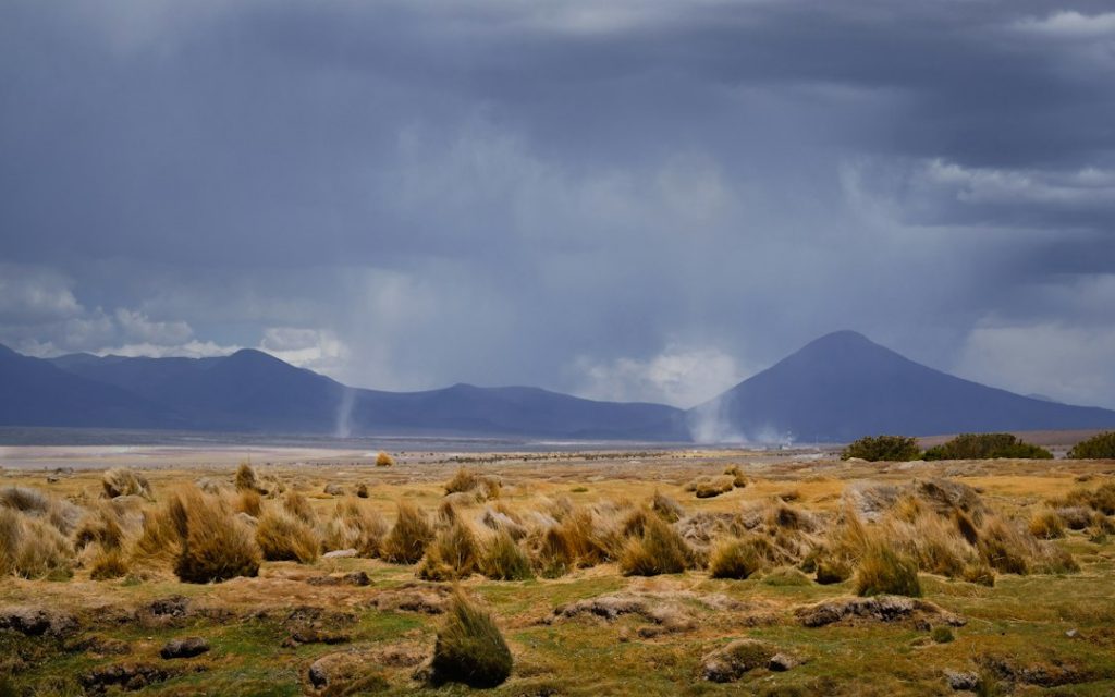 A dry, grassy plain with tufts of yellow-brown grass and rocks, set against distant dark mountains under a cloudy sky. Rain showers or mist fall from the clouds in streaks over the mountains.