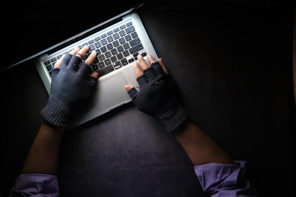 A person wearing fingerless gloves types on a laptop keyboard, viewed from above against a dark background.