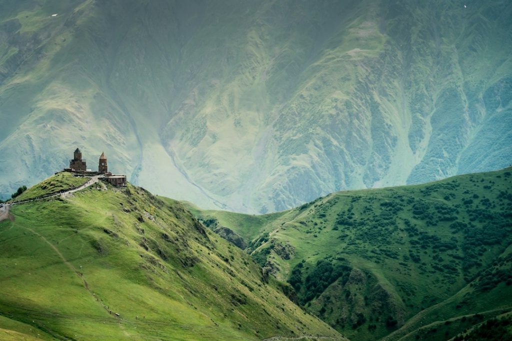A small stone church sits atop a lush, green hill, with dramatic, misty mountains rising in the background. The scene is peaceful and remote, surrounded by rolling grassy slopes.