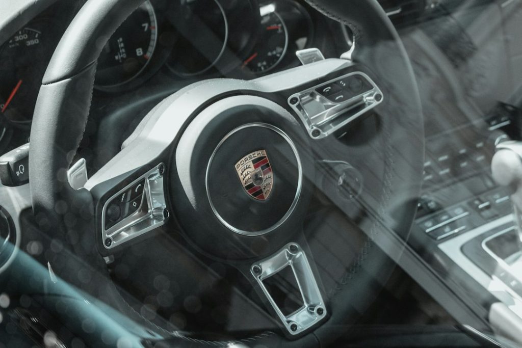 Close-up view of a Porsche steering wheel and dashboard, photographed through the car window, showing the Porsche logo and control buttons on the wheel.