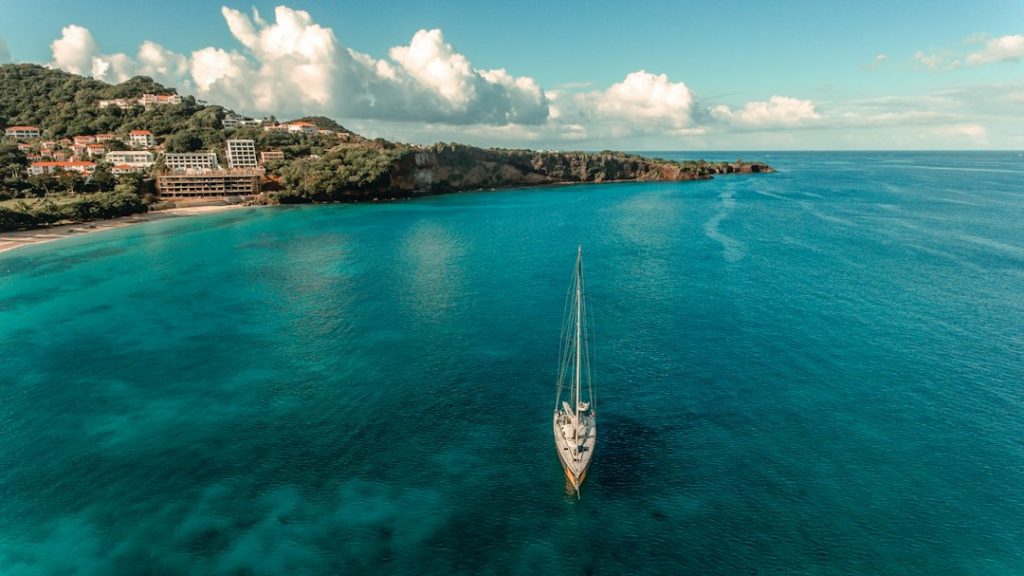 A sailboat floats on clear blue water near a lush, green coastline with scattered houses and hotels under a partly cloudy sky.