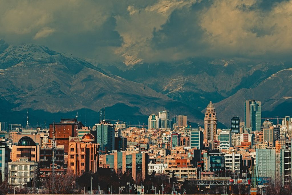 A cityscape with a mix of modern and traditional buildings set against dramatic, cloud-covered mountains in the background. The sky is moody and overcast, casting a dramatic light on the urban skyline.