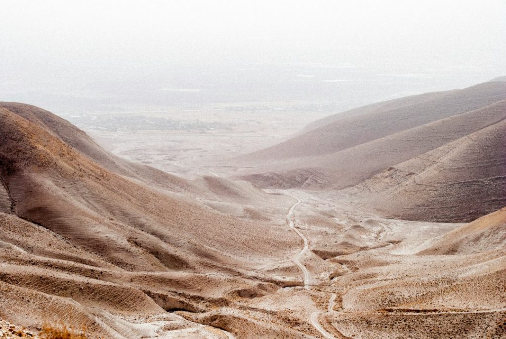 A dusty, barren landscape of rolling brown hills and valleys under a hazy sky, with faint tracks winding through the terrain. Sparse vegetation is visible, creating a desolate, arid scene.