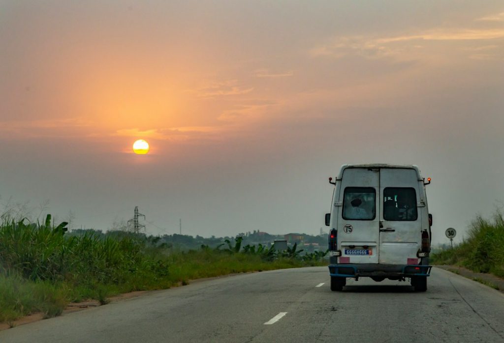 A white van drives down a rural road at sunset, with the sun low in the sky and grass lining both sides of the road. The sky is partly cloudy, and a speed limit sign is visible on the right.