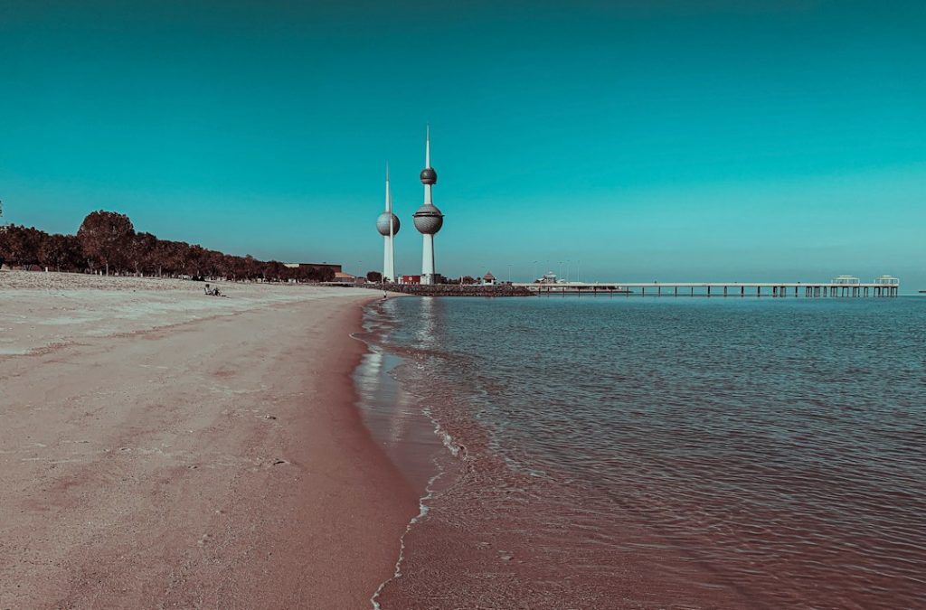 A sandy beach with calm waves, trees in the distance, and the iconic Kuwait Towers rising on the horizon under a clear blue sky. A long pier extends into the sea on the right.