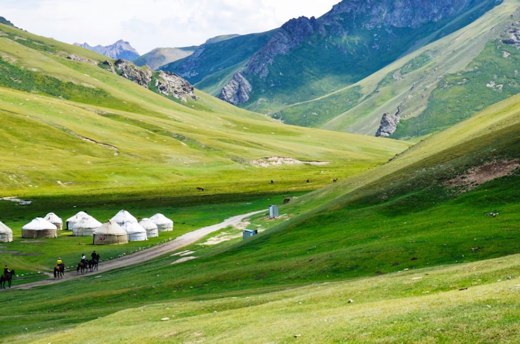 A group of yurts is set up on a bright green grassy valley, surrounded by rolling hills and mountains. Several people on horseback travel along a dirt path near the yurts under a partly cloudy sky.