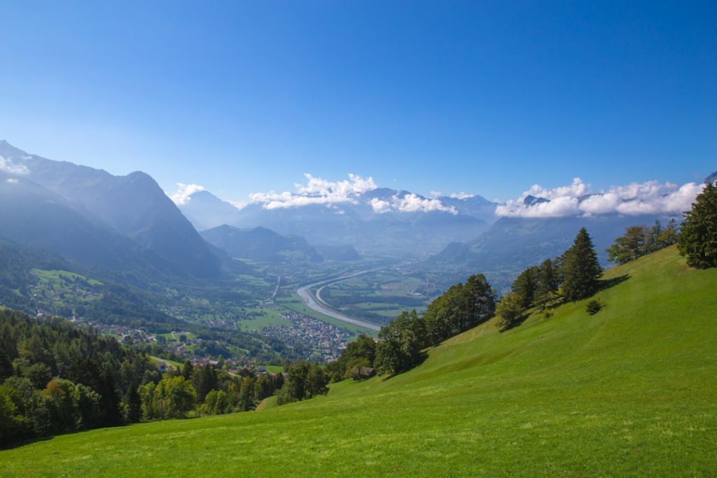 A lush green hillside overlooks a winding river and a scenic valley, with scattered towns below. Tall mountains rise in the background under a clear blue sky with some fluffy clouds.