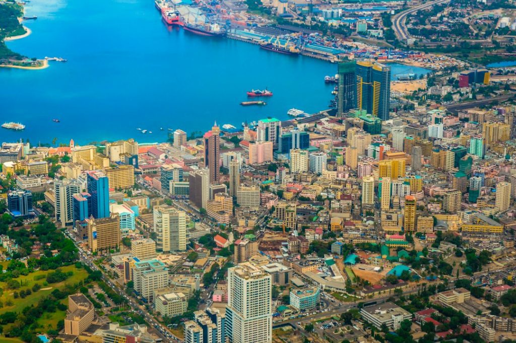 Aerial view of a vibrant coastal city with numerous colourful high-rise buildings, busy streets, green spaces, and a large harbour with ships docked along the blue waterfront.