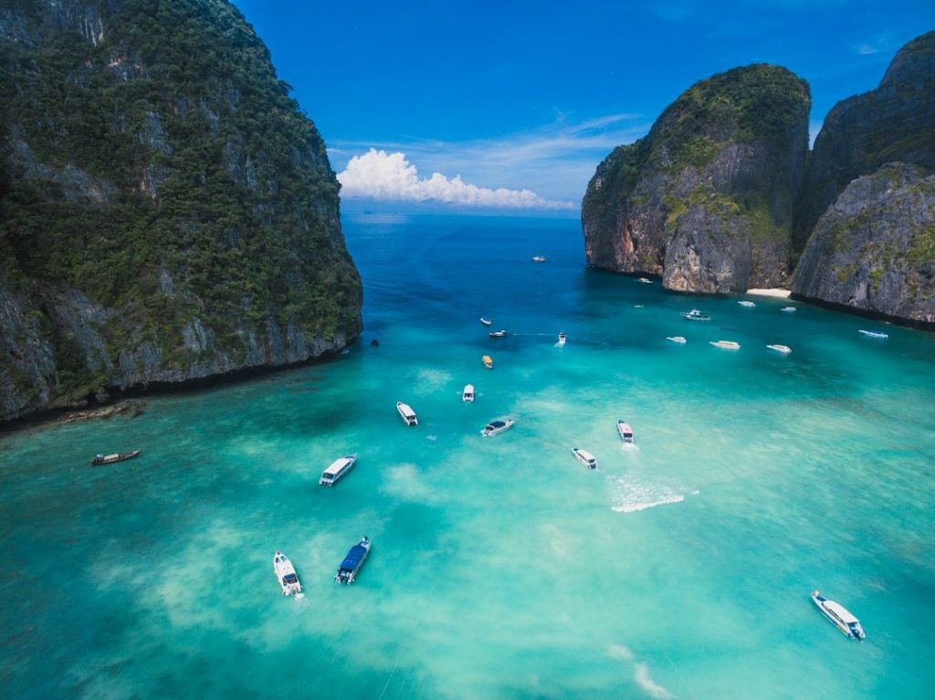 Aerial view of turquoise water with several boats near a tropical bay, surrounded by steep green cliffs and rocky islands under a bright blue sky.