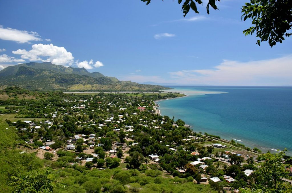 A coastal town with clusters of houses surrounded by lush greenery, next to a blue sea and bordered by mountains under a clear, sunny sky.