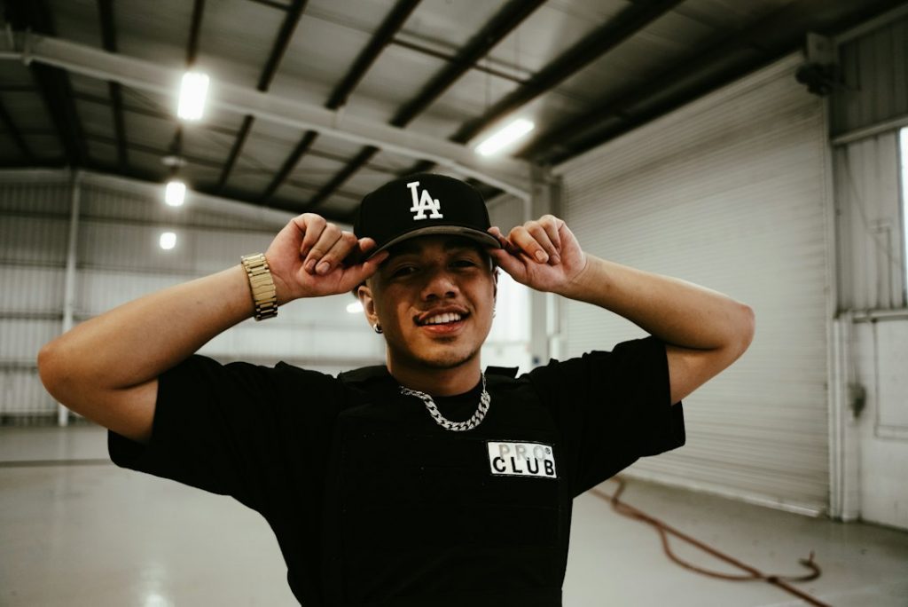 A person in a black PRO CLUB shirt and gold watch adjusts a black LA baseball cap, smiling inside a spacious, industrial warehouse with high ceilings and bright lighting.