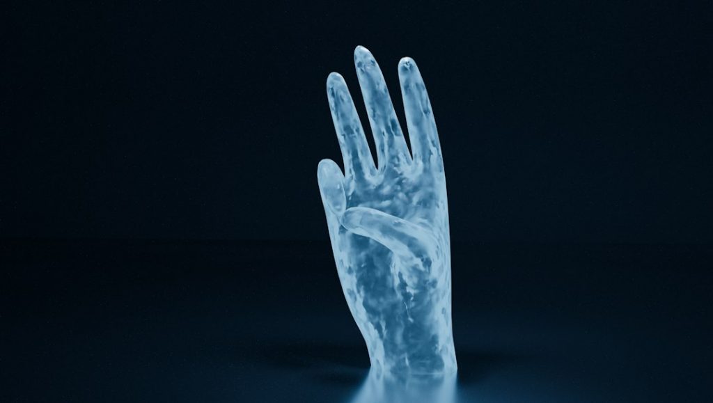 A translucent, blue-tinted hand sculpture rises from a dark, smooth surface against a black background, illuminated with soft light that highlights its semi-transparent texture.