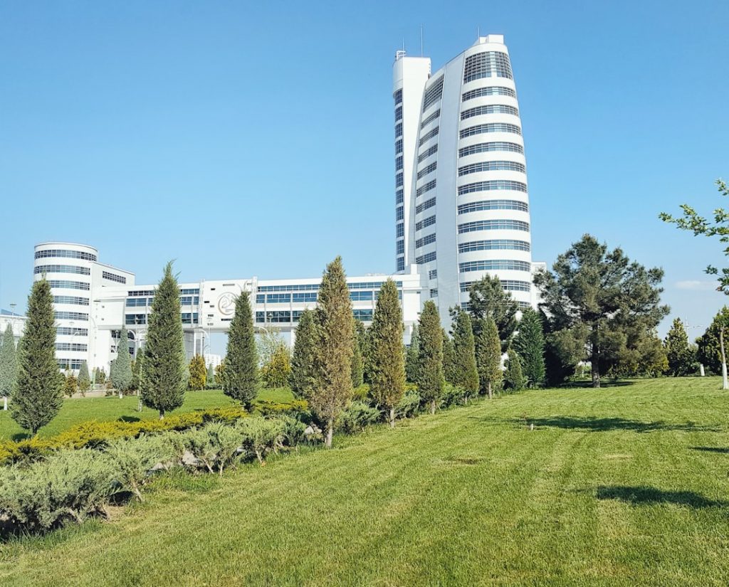 A modern white building with curved, glass-fronted towers stands behind a row of tall, green trees and a well-manicured grassy lawn under a clear blue sky.