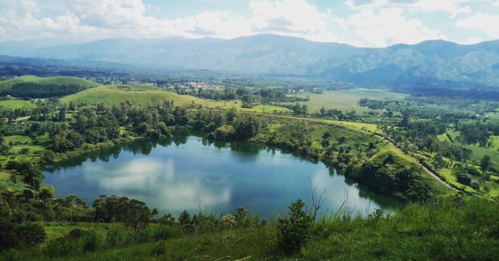 A round crater lake surrounded by lush green hills and trees, with distant mountains and a cloudy sky in the background. The water reflects the sky and some clouds.