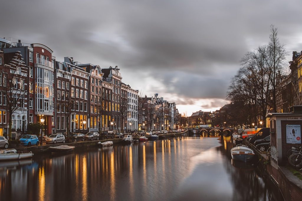 A canal in Amsterdam at dusk, with historic buildings and parked cars lining the water. Boats are moored along the canal, and the sky is cloudy with soft evening light reflecting on the water.