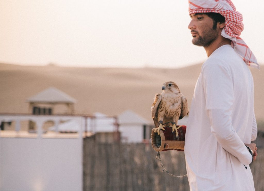 A man wearing a white thobe and red-chequered headscarf stands in a desert setting, holding a falcon on his gloved hand. Traditional Middle Eastern buildings and sand dunes are visible in the background.