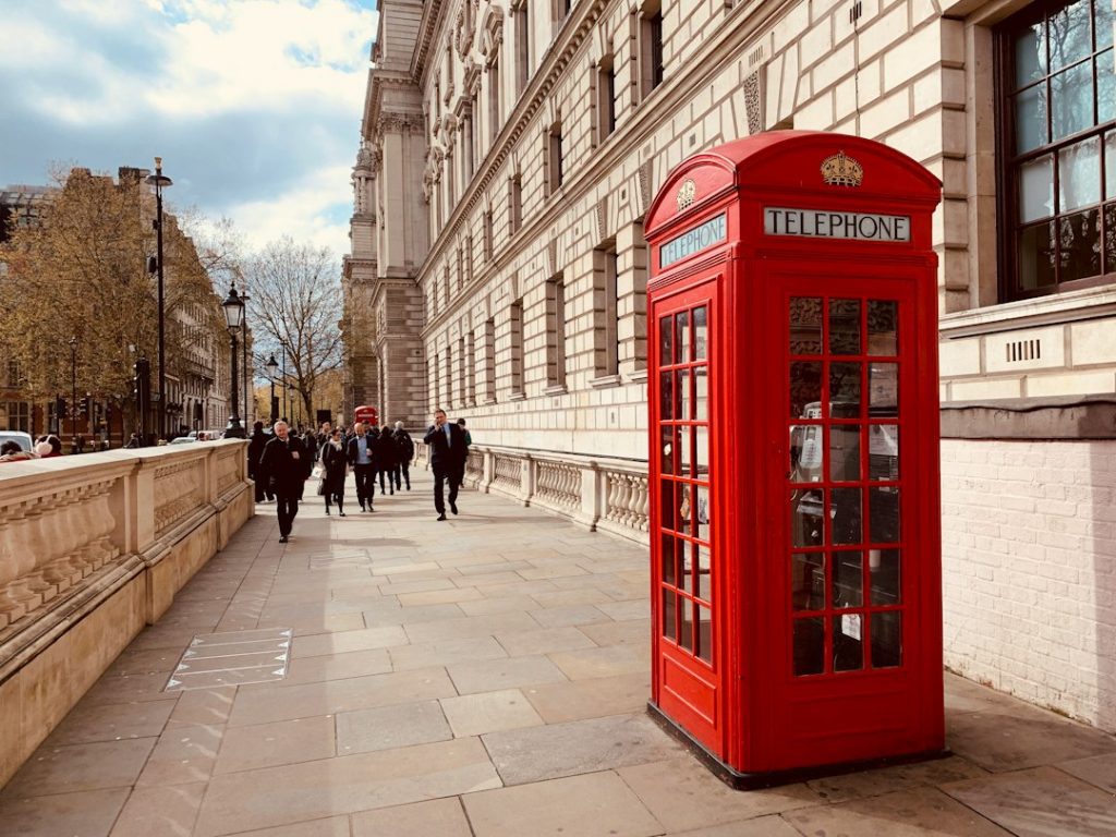 A classic red British telephone box stands on a wide pavement beside a historic stone building, with people walking in the background under a partly cloudy sky.