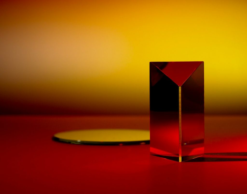 A glass prism stands upright on a red surface with a round glass disc lying flat nearby. The background fades from deep red at the bottom to bright yellow at the top, creating a vibrant, abstract scene.