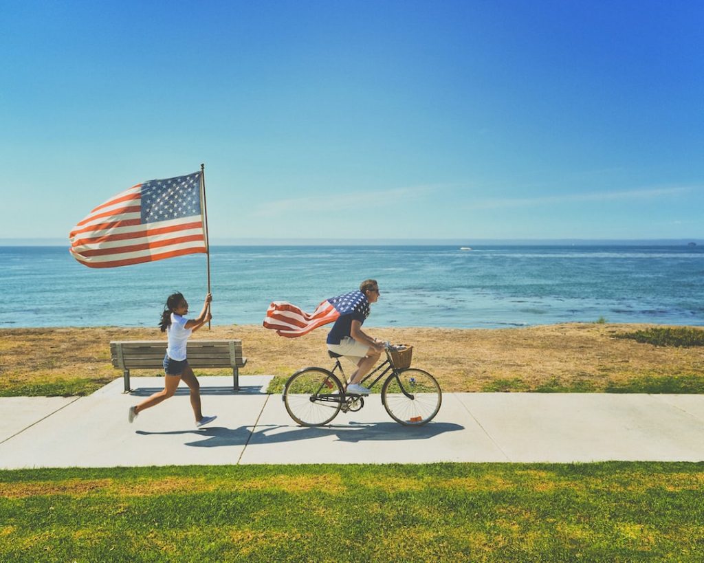 A man rides a bicycle along a seaside path, draped in an American flag, whilst a girl runs behind him holding another large American flag. The sea and clear blue sky are in the background.
