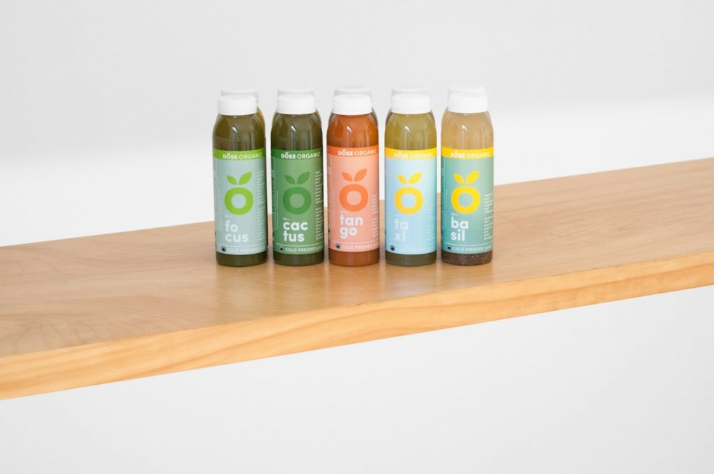 Six bottles of juice with colourful labels reading focus, cactus, tango, taxi, and basil are lined up on a light wooden shelf against a plain white background.