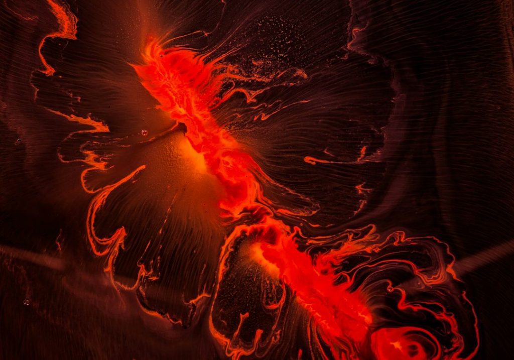 Abstract image featuring bright, fiery red and orange liquid patterns spreading across a dark background, resembling molten lava or energy waves radiating from a central glowing area.