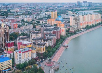 Aerial view of a Kazakhstani city with colourful high-rise buildings along a riverbank, tree-lined streets, and modern architecture. A promenade runs beside the river, with more buildings visible in the distant background.